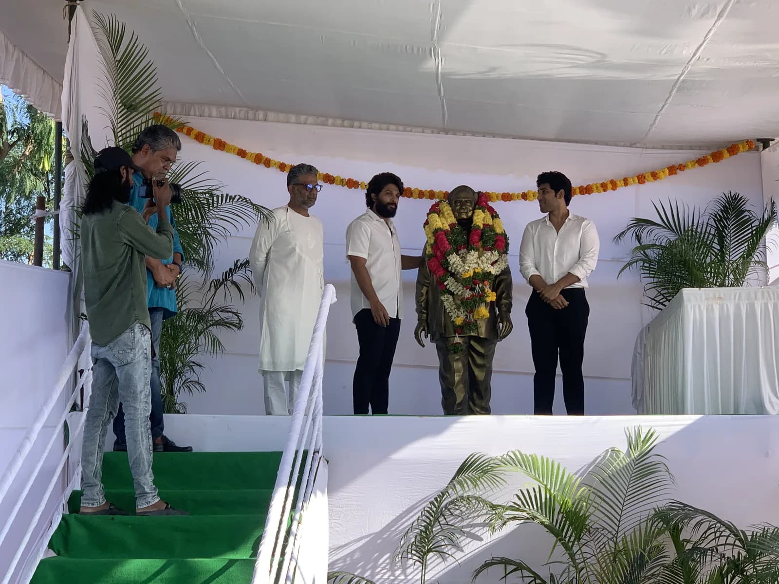 Guests gathered around the garlanded statue under the canopy, one photographing the ceremony