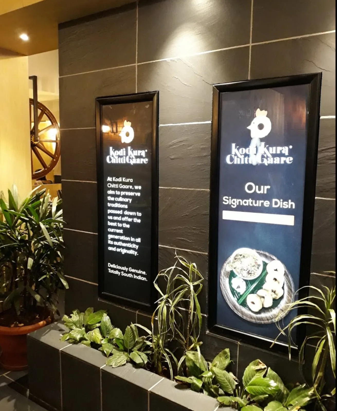 A lit signature-dish panel beside a planted bed at the entrance