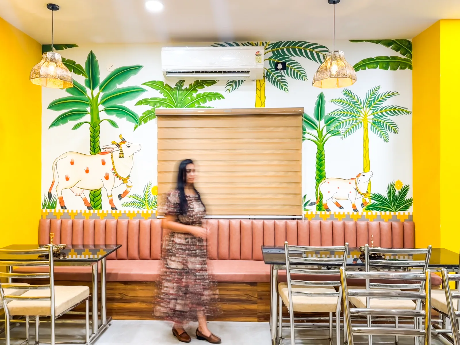 A dining room with a painted palm and cattle mural above banquette seating, a guest standing in front